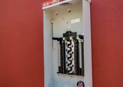 An open electrical panel mounted on a red wall with no cover, exposing the circuit breakers and wiring.