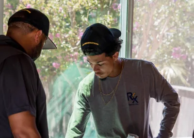 Two men discussing a set of blueprints indoors near a window with a flowering tree visible outside.