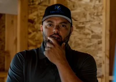 A man in a black cap and polo shirt standing indoors with a contemplative expression, hand on chin, with wooden beams in the background.